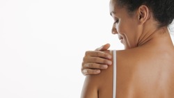 Horizontal close-up of slim African American woman strokes her shoulder standing with her back on white background | Skin moisturizing and body care concept