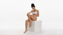 Young slim pretty African American woman with curly hair in bun in white underwear strokes smooth leg sitting on white cube platform on white background | Skin care concept
