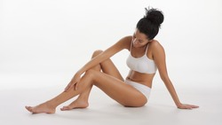 Horizontal long shot of African American woman in white underwear strokes her leg sitting on the floor on white background | Smooth legs and unwanted hair removal concept