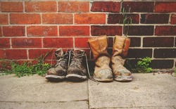 Two pairs of old boots in front of a  brick wall.The boots are heavily worn steel toe cap safety boots.