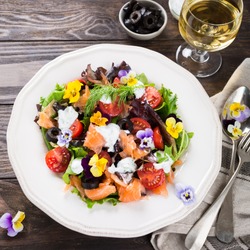 Fresh salad with smoked salmon, black olives, cherry tomatoes and edible flowers on wooden background.