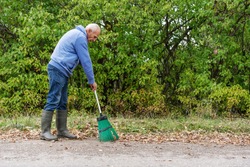 old grey haired gardener man sweeping dry foliage on asphalt road side view