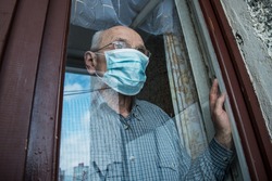 Sad aged man in face mask looking through glazing window surface. Concept lockdown during pandemic.