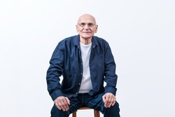 Old man in eyeglasses wearing blue shirt and jeans seated on wooden chair isolated on white background in studio