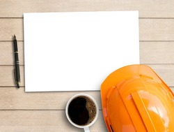 Blank notepad,orange safety helmet and cup of coffee on brown wood table background. Top view with copy space for text or any design. White paper on table wood show paper concept