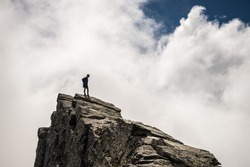 Hiker standing on steep rocky mountain summit without protection. Dramatic cloudy sky. Concept of reaching the goal and conquering the success. Contrasted, silhouette like.