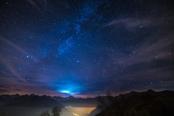 The wonderful starry sky on Christmas time and the majestic high mountain range of the Italian French Alps, with glowing villages below and moonlight behind the clouds.
