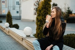 A beautiful girl with long hair with coffee sitting on a bench and looking away.