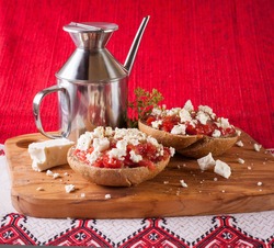 Dakos, Crete meze with barley rusk paximadia with tomato, feta or myzithra cheese and oregano and olive oil with metal oilcan on the olive wood cutting board and white ornament cloth horizontal