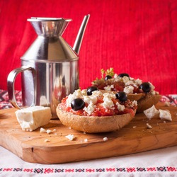 Dakos, Crete meze with barley rusk paximadia with tomato, feta cheese, black olive and oregano and olive oil with metal oilcan on the olive wood cutting board with red and white ornament cloth quader