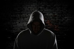 An evil looking man with a hidden face and wearing a hoodie stands intimidatingly against an ally wall stalking his next victim.