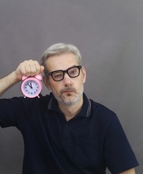 Middle age men with gray hair holding pink alarm clock on gray background. Think about time, aging process and deadline concept.