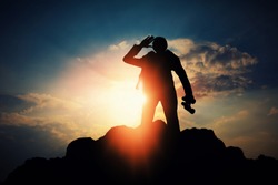 silhouette of businessman with binocular for searching a opportunity on top of mountain against sun light 