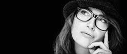 Monochrome close-up cropped portrait of a beautiful pensive woman in stylish glasses wearing a black hat looking up to the side with hand to chin in a concept of eye care and ophthalmology