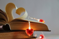 On a light background, a stack of books with dark covers. Book pages in the shape of a heart. A luminous garland with red glass or plastic hearts lies on the book. Romantic evening. Valentine's Day.