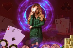 Blonde girl in green dress has covered her eye by two aces, posing on colorful background with neon circles, cards, chips. Poker, casino. Close-up
