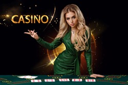 Lady in green dress is showing inscription casino, leaning on playing table with cards on it, posing on black background. Poker, casino. Copy space