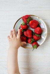 Hand of a child with a strawberry on a rustic background, a plate of strawberries. The concept of summer healthy eating. View from above, apartment.