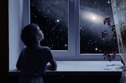 A little boy is standing near the window and looking outside, imagining boundless space with myriad of stars