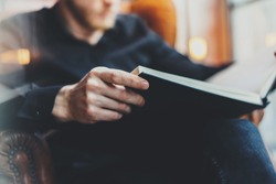 Closeup view of male hands holding book.Young bearded man relaxing at home while sitting in vintage chair.Selective focus on hand,blurred background.Horizontal, film effect