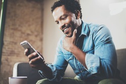 Happy bearded African businessman using phone while sitting on sofa at his modern home.Concept of young people working mobile devices.Blurred background