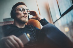 Portrait handsome bearded man wearing glasses,headphones listening to music at modern home.Guy sitting in vintage chair,holding smartphone and relaxing.Panoramic windows background.Blurred background