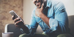 Attractive bearded African businessman using smartphone while sitting on sofa at his home.Concept of young people working mobile devices.Closeup with a selected focus.Blurred background.Wide