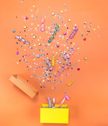 Yelow gift box with various party confetti, streamers, noisemakers and decoration on a orange background. Colorful celebration background.