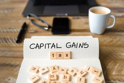 Closeup on notebook over vintage desk surface, front focus on wooden blocks with letters making Capital Gains Tax text. Business concept image with office tools and coffee cup in background