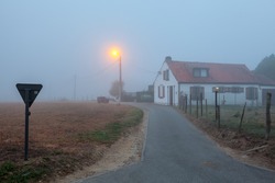 Next to the road longest a field there is a house in the fog