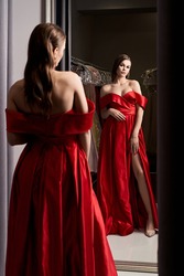 Young beautiful woman wearing an off-the-shoulder full-length crimson red satin slit prom ball gown. Model looking in mirror.