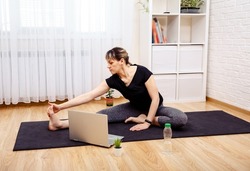Photo of young concentrated woman doing yoga exercises using laptop while sitting on floor at home. Yoga exercises at home. Self-isolation is beneficial, entertainment and education on the Internet.
