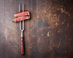 Slices of beef steak on meat fork on dark wooden background