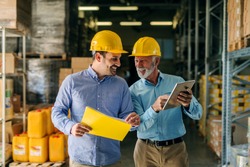 Two successful smiling business man walking through big warehouse with helmets on their heads. Older man is holding digital tablet and shoving younger one some documents.