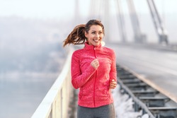 Happy Caucasian woman in sportswear and with ponytail running on the bridge at winter. Winter fitness concept.