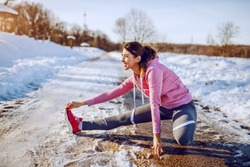 Side view of beautiful caucasian woman in sportswear and with ponytail stretching leg on country road. Winter time. Outdoor fitness concept.