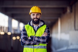 Cute Caucasian bearded construction worker with safety helmet on head in vest standing with arms crossed at construction site and looking at camera.