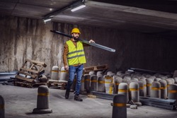 Full length of handsome caucasian bearded worker in vest and helmet on head carrying metal rod on shoulder. Tunnel in construction process interior.