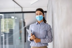 Young businesswoman with face mask standing in corporate firm with tablet in hands during corona virus.