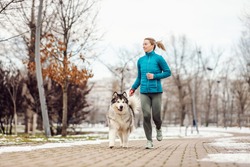 Sportswoman running in park with her dog on a leash on a cold winter day. Healthy lifestyle, cold weather, snow, city life