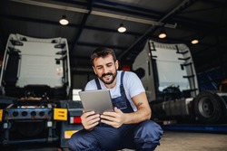 Smiling tattooed bearded blue collar worker in overalls using tablet to check on delivery while crouching in garage of import and export firm. In background are trucks.