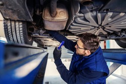 Technical inspection of the car. Car service in workshop. A man in a blue uniform stands under a car in a garage and checks the condition of the brakes on car tires. Troubleshooting motorcar