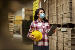 A woman in a plaid shirt and a protective helmet under her arm passes a digital tablet through the warehouse and checks the condition of the raw materials. She has a mask due to the corona situation
