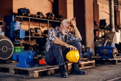 An old, tired bearded factory worker in overalls is sitting on the pallet and taking a break from hard work. He is wiping sweat from his forehead.