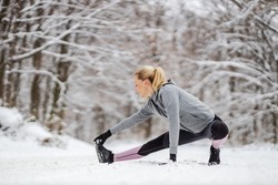 Flexible fit sportswoman crouching in nature and doing warmup and stretching exercises at snowy winter day. Sportswear, exercises in nature, winter fitness