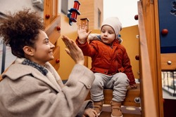 Kindergarten child giving high five to nursery governess on a playground. A cute little boy is sitting in a castle on a playground and giving a high five to his nursery governess.