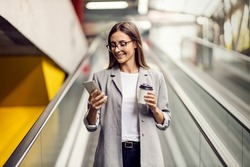 A happy, elegant businesswoman descending the escalator and holding takeaway coffee while reading important messages. A woman on the escalator with a phone