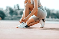 Sporty female tying shoelaces in training. A woman in a sports outfit squats and ties a shoelace. Gray wardrobe and white sneakers at the outdoor training stadium, close shot of a female body