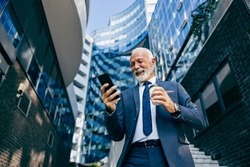 An old executive reading message on the phone at the business center. Low angle view of a happy senior businessman standing at the business center and reading a message on the phone.
