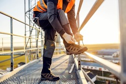 An industry worker tying shoelace on work shoes while standing on metal construction.